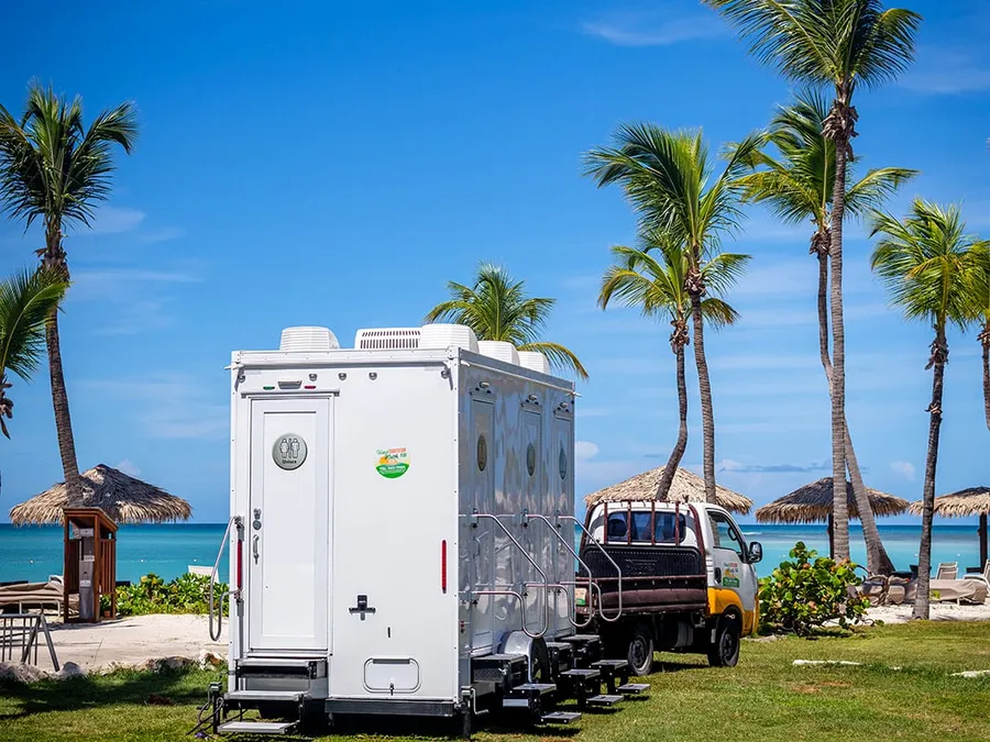 Island Sanitation Plus AC restroom trailer and truck set up on the beach at a resort, palm trees and ocean behind